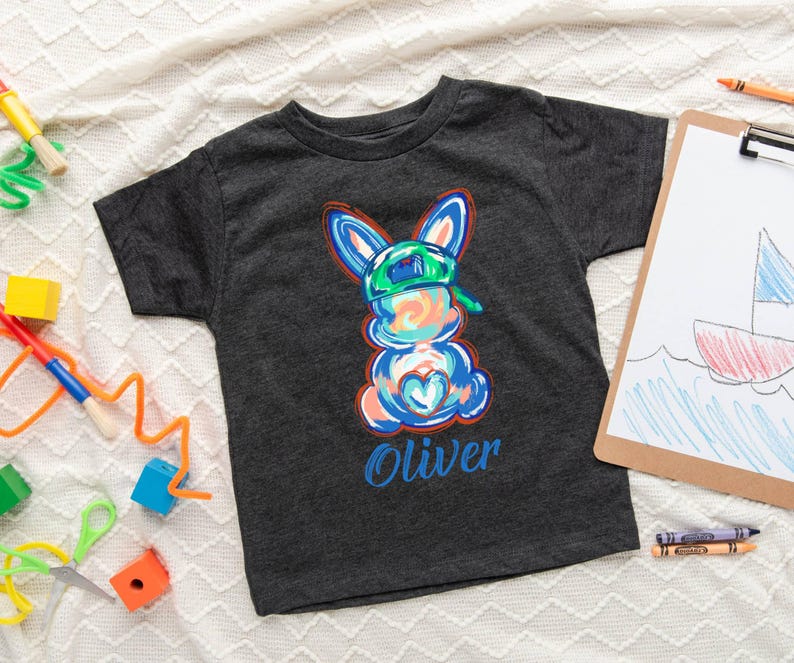 May include: A dark grey t-shirt with a colorful cartoon bunny wearing a green hat. The bunny has a blue heart on its chest and the name "Oliver" is printed below it.