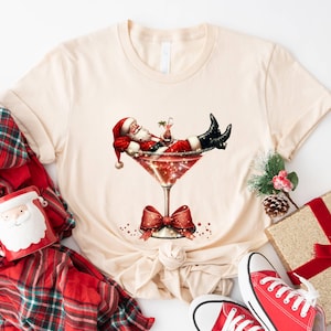 May include: A beige t-shirt with a graphic of Santa Claus relaxing in a martini glass. The glass is decorated with glitter and a red bow. The graphic is surrounded by red and white polka dots.