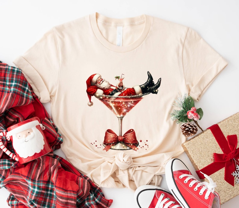 May include: A beige t-shirt with a graphic of Santa Claus relaxing in a martini glass. The glass is decorated with glitter and a red bow. The graphic is surrounded by red and white polka dots.