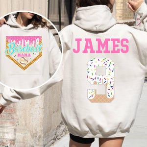 K&ouml;nnte beinhalten: Wei&szlig;er Hoodie mit einem rosafarbenen Baseball-Motiv auf der Vorderseite und dem Namen "JAMES" und der Nummer "9" auf der R&uuml;ckseite. Das Motiv zeigt ein Baseballfeld mit einem tropfenden Eiswaffel und Streuseln.