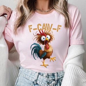 May include: Pink t-shirt featuring a cartoon rooster with wide eyes and an open beak, with the text "F-CAW-F" in gold letters. The rooster has red, brown, blue, and yellow coloring.