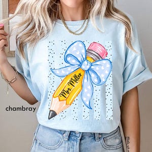 May include: A light blue t-shirt featuring a graphic of a yellow pencil with a pink eraser and a large light blue bow with white hearts. The pencil has the text Mrs. Miller written in black script. The background of the graphic has black dots.
