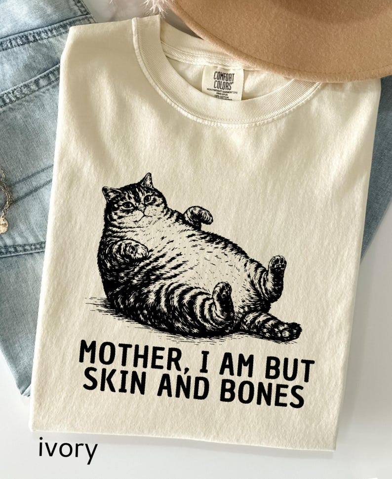 以下が含まれることがあります： アイボリー色のTシャツ。黒と白で描かれた、仰向けに寝そべる猫のイラストが特徴。猫の下には「MOTHER, I AM BUT SKIN AND BONES」の文字。柔らかい素材で作られています。