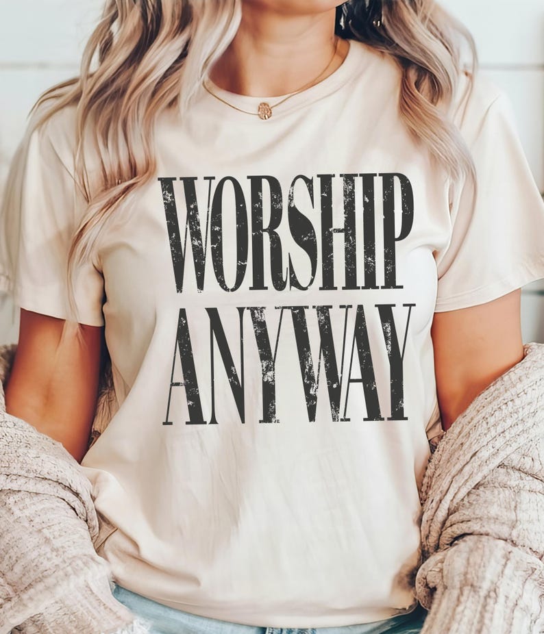May include: A beige t-shirt with the words "WORSHIP ANYWAY" printed in a distressed black font. The shirt is short-sleeved and has a crew neck. The person is wearing a gold necklace and a beige cardigan.