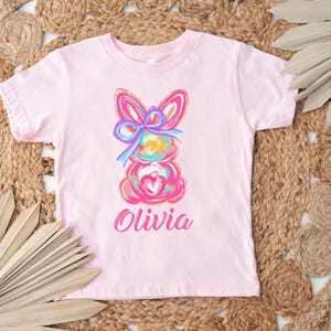 May include: Pink t-shirt with a colorful abstract bunny design and the name "Olivia" printed below.
