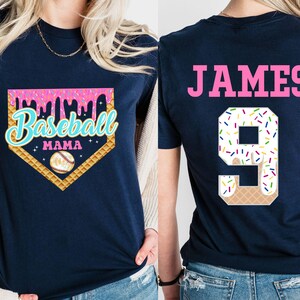 May include: Two navy blue t-shirts. The front shirt has a pink, white, and blue baseball-themed graphic with the text "Baseball Mama". The back shirt has the name "JAMES" and the number "9" in pink with a white and colorful ice cream cone design.