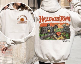 May include: A cream-coloured hoodie with a Halloween theme. The front has a circular design with "Halloweentown University" and a jack-o'-lantern. The back shows a colourful town scene with a large pumpkin and the text "Halloweentown EST. 1998".