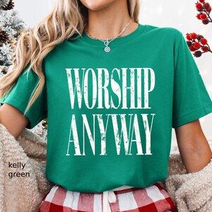 May include: Kelly green t-shirt with the words "WORSHIP ANYWAY" in distressed white lettering. The shirt is a crew neck style. The model is wearing red and white plaid pajama pants.