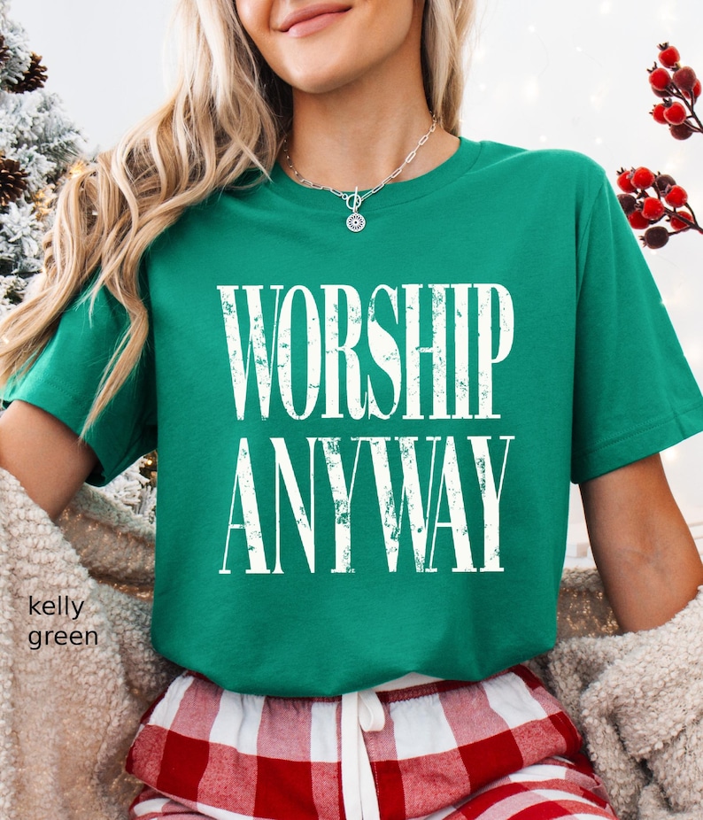 May include: Kelly green t-shirt with the words "WORSHIP ANYWAY" in distressed white lettering. The shirt is a crew neck style. The model is wearing red and white plaid pajama pants.