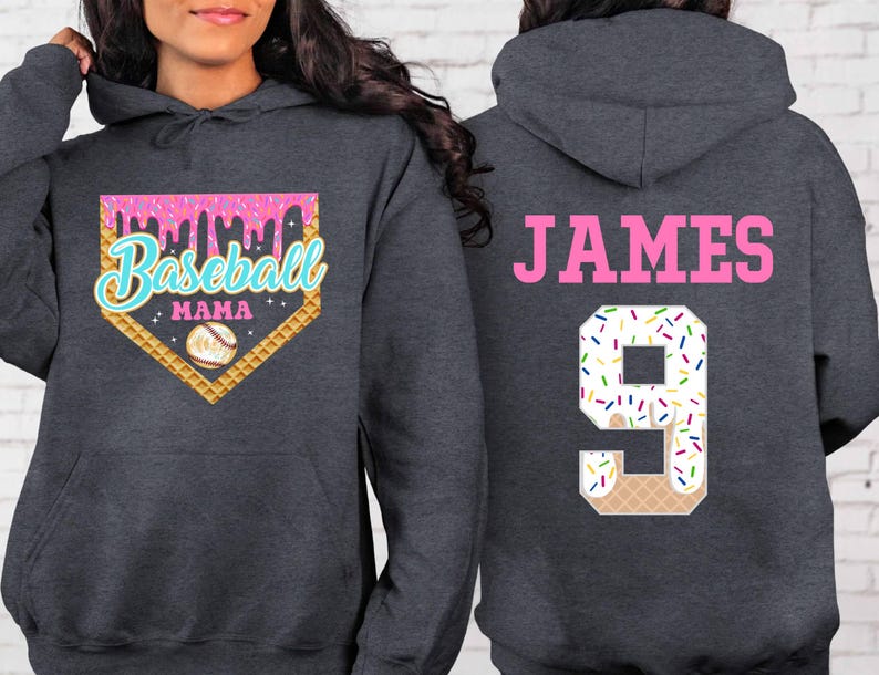 K&ouml;nnte beinhalten: Ein dunkelgrauer Kapuzenpullover mit einem pinkfarbenen, blauen und wei&szlig;en Baseball-Motiv auf der Vorderseite. Das Motiv lautet "Baseball Mama" und zeigt einen Baseball in einem Waffelh&ouml;rnchen. Die R&uuml;ckseite des Pullovers hat ein pinkfarbenes "JAMES" und eine wei&szlig;e Nummer "9" mit Streuseln und einem Waffelh&ouml;rnchen-Design.