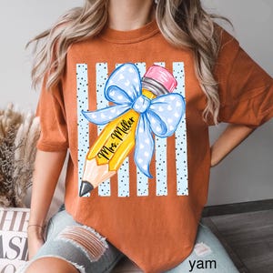 May include: A burnt orange t-shirt featuring a graphic of a yellow pencil with a pink eraser and a large light blue bow with white hearts. The pencil is printed with the text Mrs. Miller and is set against a background of light blue vertical stripes.
