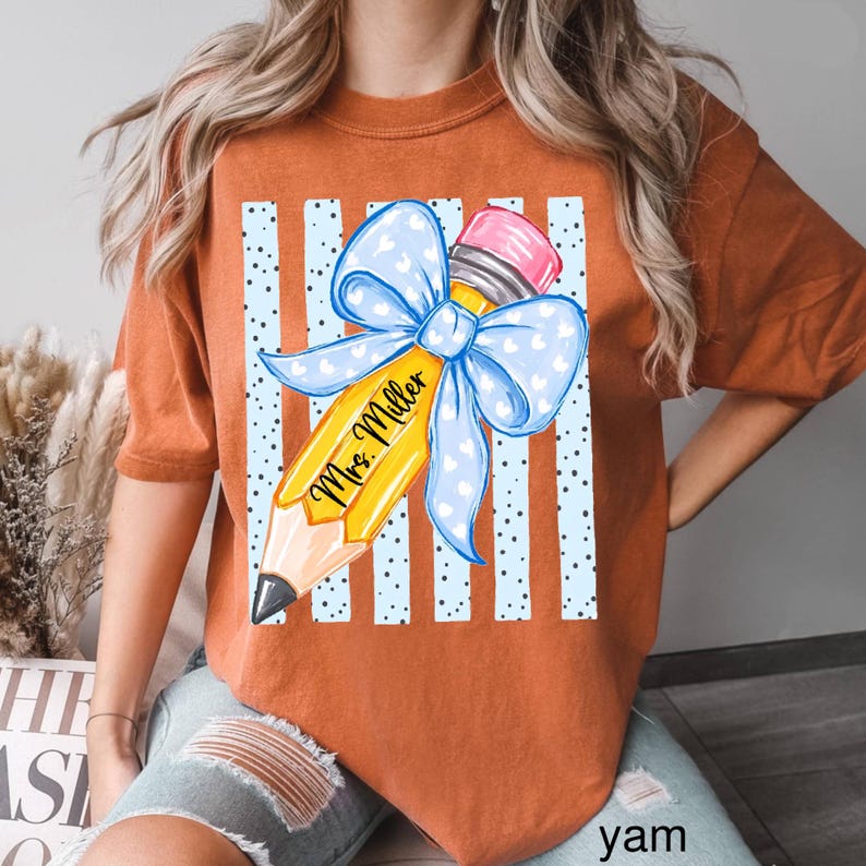 May include: A burnt orange t-shirt featuring a graphic of a yellow pencil with a pink eraser and a large light blue bow with white hearts. The pencil is printed with the text Mrs. Miller and is set against a background of light blue vertical stripes.