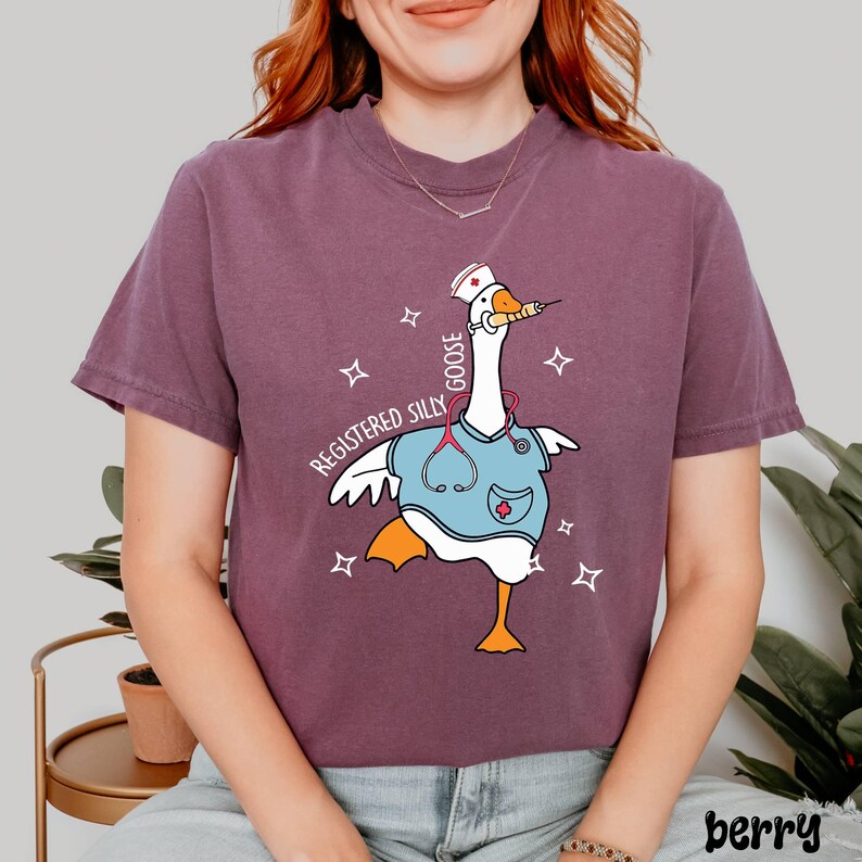 K&ouml;nnte beinhalten: Ein lila T-Shirt mit einer Cartoon-Gans, die eine Krankenschwesteruniform und einen Hut tr&auml;gt. Der Text auf dem Shirt lautet "Registered Silly Goose".