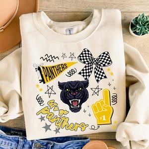 May include: Cream-colored sweatshirt featuring a black panther graphic, a checkered bow, and the text "Let's go Panthers." Includes a yellow pennant with "Panthers" and a foam finger with the number one.