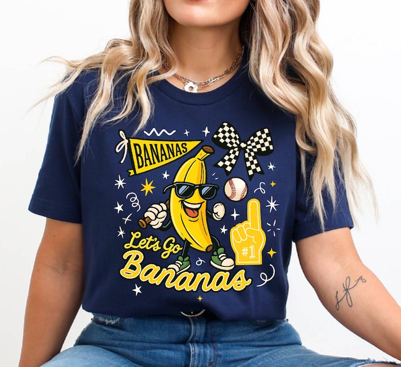May include: Navy blue t-shirt with a playful graphic featuring a smiling banana wearing sunglasses, a baseball, and a foam finger. The text "Let's Go Bananas" is displayed in a whimsical font, with stars and other decorative elements.