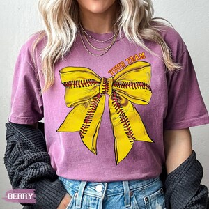 May include: Berry-colored t-shirt featuring a large yellow softball bow graphic with red stitching. The text "YOUR TEAM" is arched above the bow. The shirt is a comfortable fit.