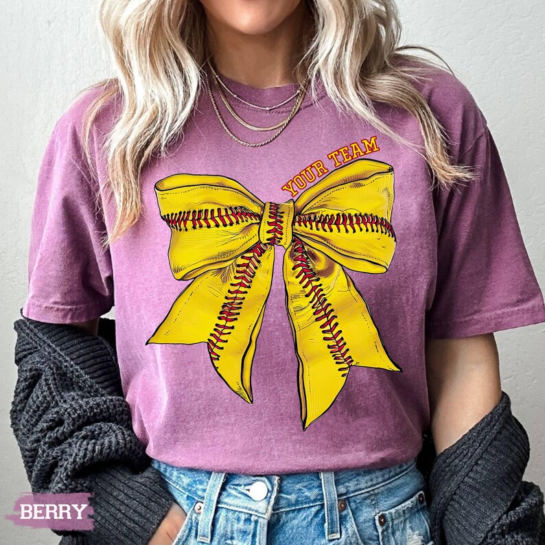 May include: Berry-colored t-shirt featuring a large yellow softball bow graphic with red stitching. The text "YOUR TEAM" is arched above the bow. The shirt is a comfortable fit.
