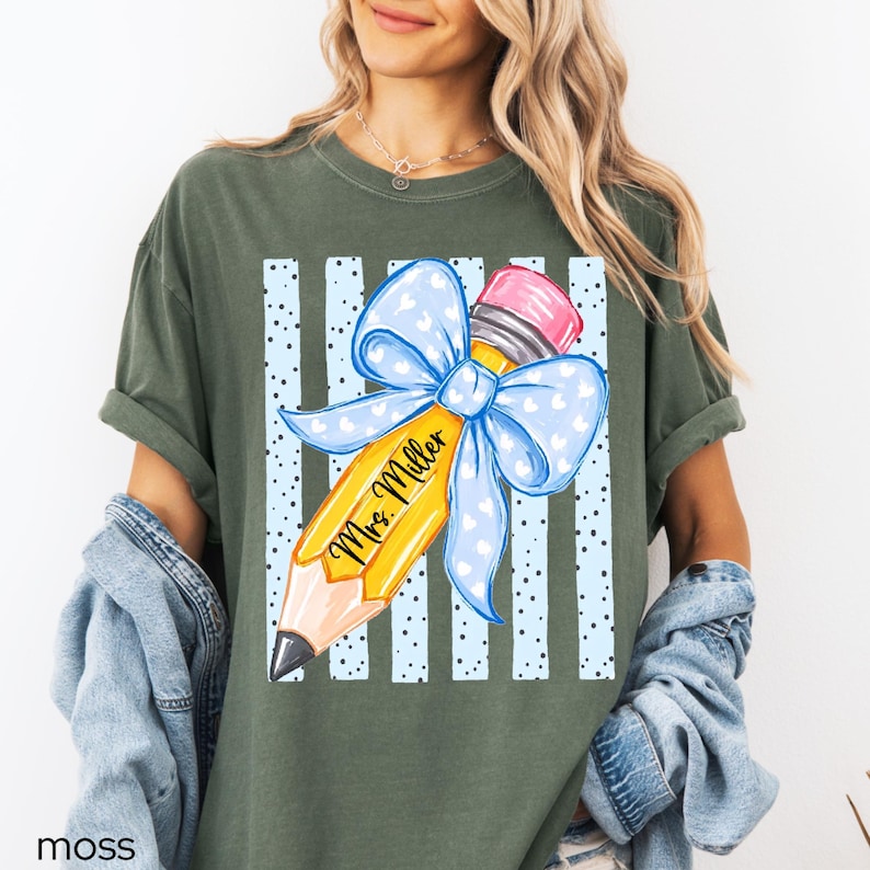 May include: A moss green t-shirt featuring a graphic of a yellow pencil with a large light blue bow tied around it. The pencil has the text Mrs. Miller written on it. Behind the pencil are vertical light blue stripes with black polka dots.