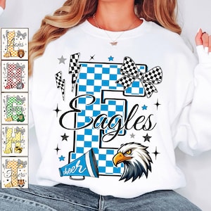 May include: White sweatshirt featuring a large blue and white checkered "E" with the word "Eagles" in script. The design includes a bow, stars, lightning bolts, a megaphone with "cheer", and an eagle head. Additional team logos are displayed on the left.