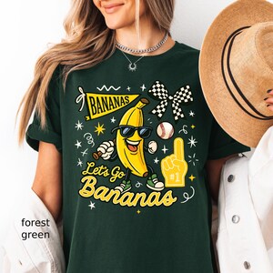 May include: Forest green t-shirt with a graphic of a smiling banana wearing sunglasses, a baseball, and the text "Let's Go Bananas". The design includes a checkered bow and a foam finger.