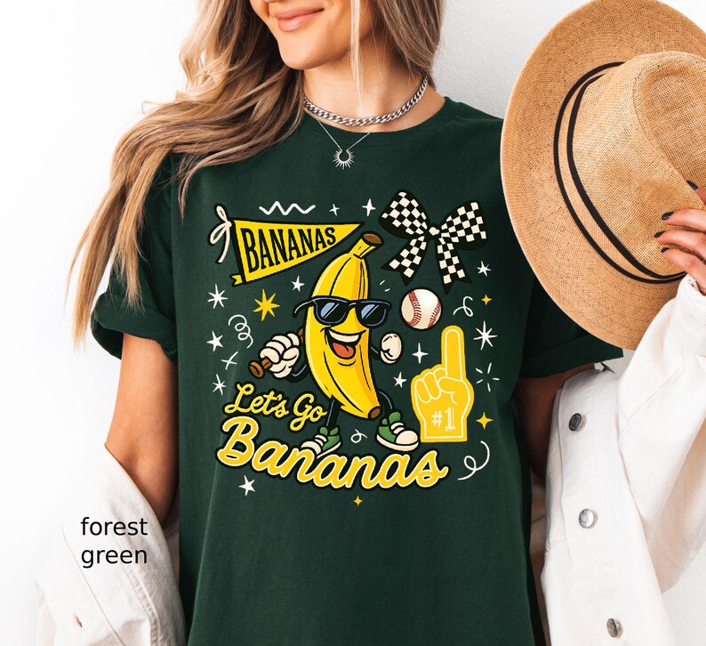 May include: Forest green t-shirt with a graphic of a smiling banana wearing sunglasses, a baseball, and the text "Let's Go Bananas". The design includes a checkered bow and a foam finger.