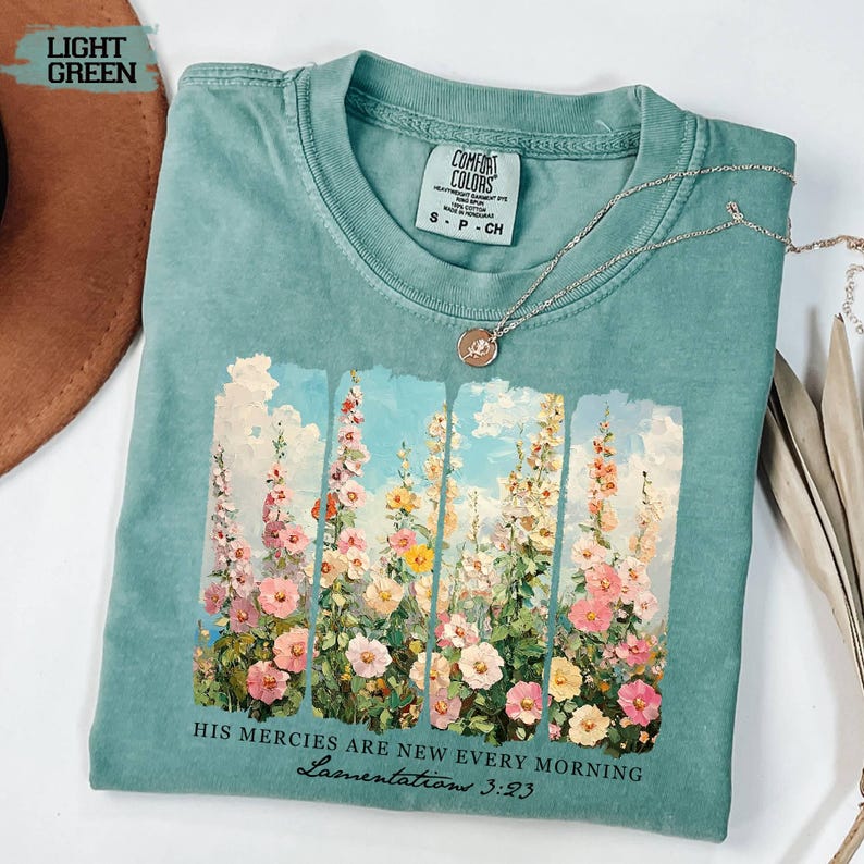 May include: Light green Comfort Colors t-shirt with a floral design featuring pink, yellow, and white flowers against a blue sky backdrop. The text "HIS MERCIES ARE NEW EVERY MORNING Lamentations 3:23" is printed below the floral design. A silver necklace is draped over the shirt.