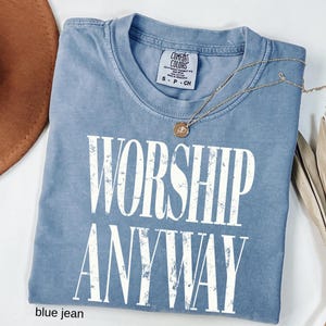May include: A blue jean Comfort Colors t-shirt with the words "WORSHIP ANYWAY" in large white letters. A gold necklace with a pendant rests on the shirt. The shirt is folded and has a tag at the neckline.