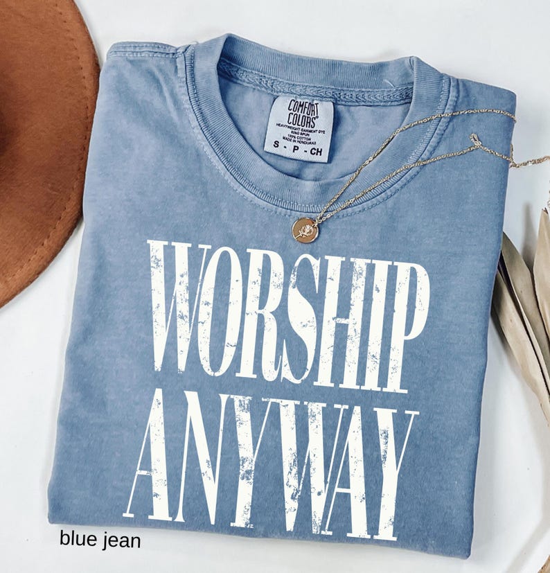 May include: A blue jean Comfort Colors t-shirt with the words "WORSHIP ANYWAY" in large white letters. A gold necklace with a pendant rests on the shirt. The shirt is folded and has a tag at the neckline.