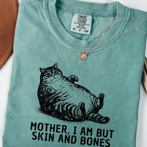 以下が含まれることがあります： 黒い猫のイラストと「MOTHER, I AM BUT SKIN AND BONES」の文字が入った、明るい緑色のTシャツ。Tシャツはクルーネックと半袖です。バラのペンダントが付いたシルバーのネックレスも確認できます。