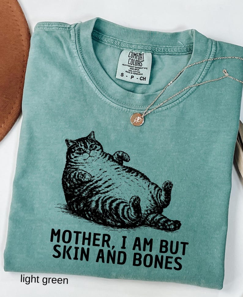 以下が含まれることがあります： 黒い猫のイラストと「MOTHER, I AM BUT SKIN AND BONES」の文字が入った、明るい緑色のTシャツ。Tシャツはクルーネックと半袖です。バラのペンダントが付いたシルバーのネックレスも確認できます。