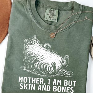 以下が含まれることがあります： リラックスした猫のグラフィックと「MOTHER, I AM BUT SKIN AND BONES」のテキストが特徴のモスグリーンのTシャツ。小さなペンダントが付いたシルバーのネックレスがシャツの上にあります。シャツには「Comfort Colors」とサイズSのラベルが付いています。