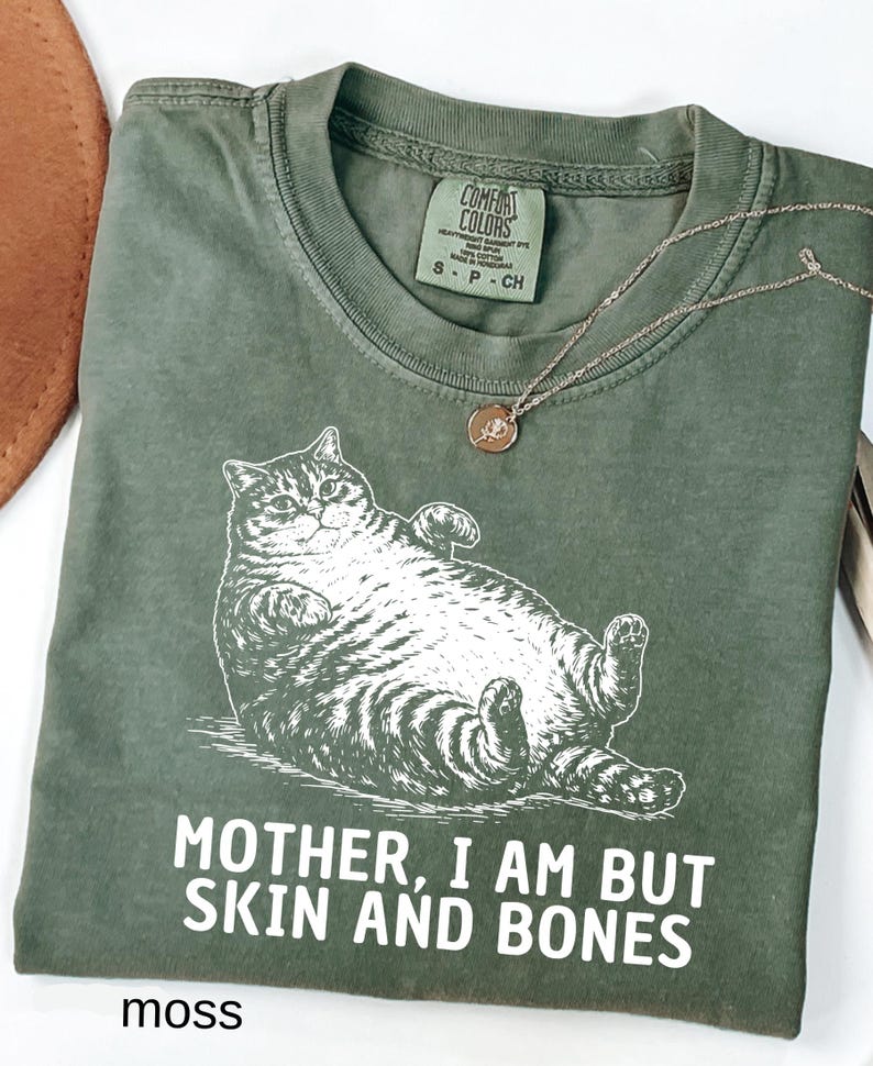 以下が含まれることがあります： リラックスした猫のグラフィックと「MOTHER, I AM BUT SKIN AND BONES」のテキストが特徴のモスグリーンのTシャツ。小さなペンダントが付いたシルバーのネックレスがシャツの上にあります。シャツには「Comfort Colors」とサイズSのラベルが付いています。