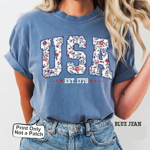May include: Blue t-shirt with the word "USA" in a floral pattern, with "EST. 1776" below. The shirt is a light blue color and has rolled-up sleeves. The text "Print Only Not a Patch" is in the bottom left corner.