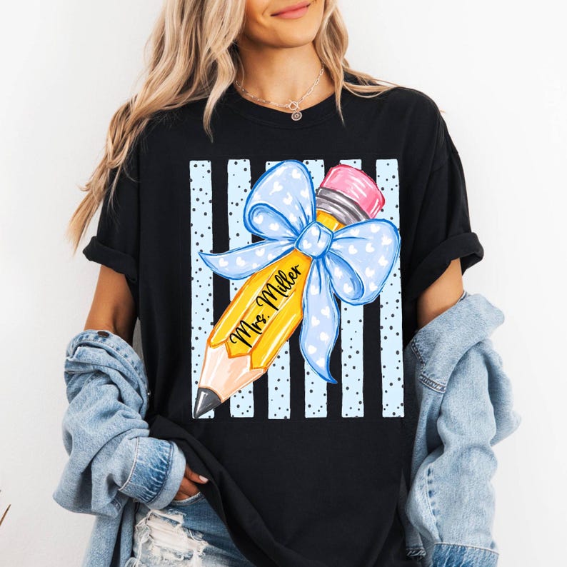 May include: A black t-shirt featuring a graphic of a yellow pencil with a pink eraser and a large light blue bow with white hearts. The pencil is inscribed with the text Mrs. Miller and is set against a background of vertical light blue stripes.