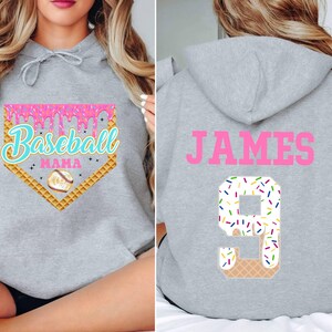 K&ouml;nnte beinhalten: Grauer Kapuzenpullover mit einem rosafarbenen Baseball-Motiv auf der Vorderseite und dem Namen "JAMES" sowie der Nummer "9" mit einem rosafarbenen und wei&szlig;en Zuckerguss auf der R&uuml;ckseite.