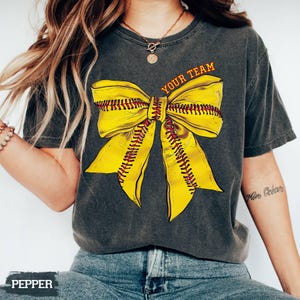 May include: Dark gray t-shirt featuring a large yellow softball bow design with red stitching. The text "YOUR TEAM" is printed above the bow in orange. The shirt is a casual style.