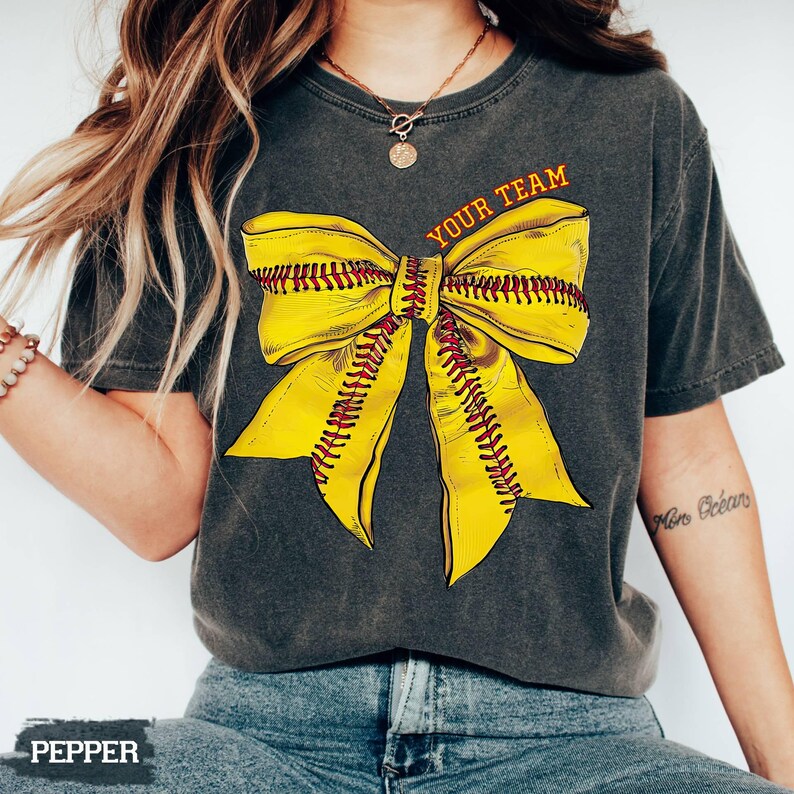 May include: Dark gray t-shirt featuring a large yellow softball bow design with red stitching. The text "YOUR TEAM" is printed above the bow in orange. The shirt is a casual style.