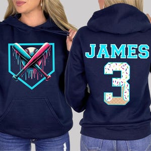 May include: Navy blue hoodie featuring a baseball-themed graphic. The front displays crossed bats and a baseball within a home plate outline. The back has the name "JAMES" and the number "3" with a sprinkle and ice cream cone design.
