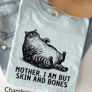 以下が含まれることがあります： 猫の白黒イラストがプリントされた水色のTシャツ。「MOTHER, I AM BUT SKIN AND BONES」の文字が猫の下にプリントされています。下部には「Chambray」の文字がプリントされています。