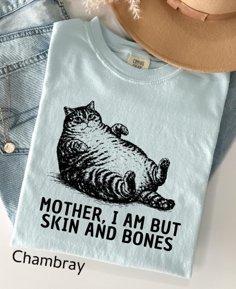 以下が含まれることがあります： 猫の白黒イラストがプリントされた水色のTシャツ。「MOTHER, I AM BUT SKIN AND BONES」の文字が猫の下にプリントされています。下部には「Chambray」の文字がプリントされています。