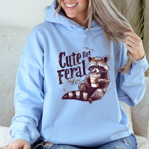 May include: Light blue hoodie with a raccoon graphic and the text "Cute But Feral". The raccoon is holding a cup of coffee.