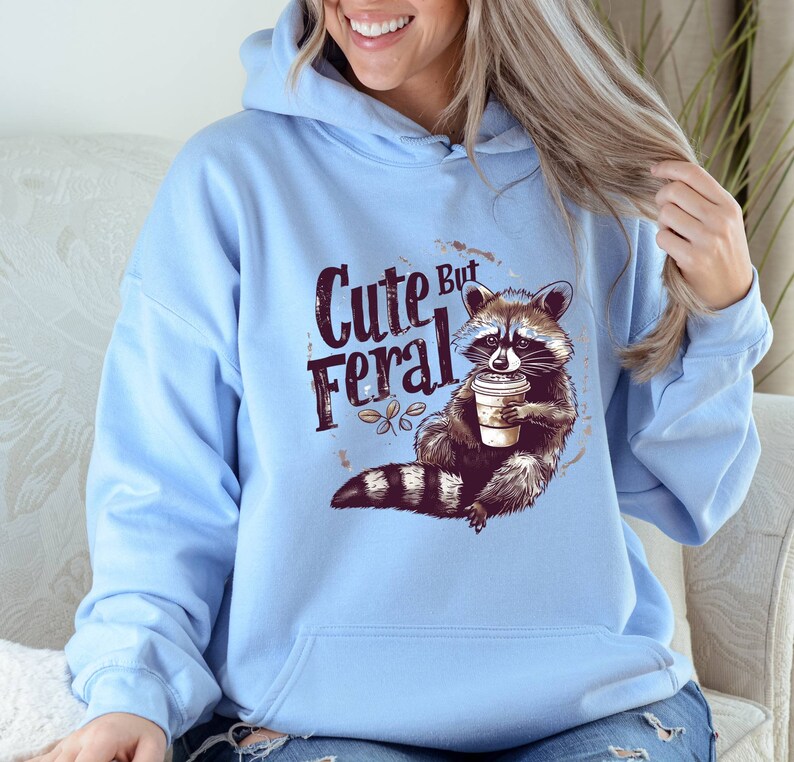May include: Light blue hoodie with a raccoon graphic and the text "Cute But Feral". The raccoon is holding a cup of coffee.