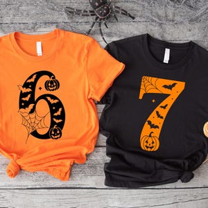 May include: Two short-sleeved Halloween-themed t-shirts. One is orange with a black number six design featuring bats, a spider web, and pumpkins. The other is black with an orange number seven design with bats and a pumpkin.