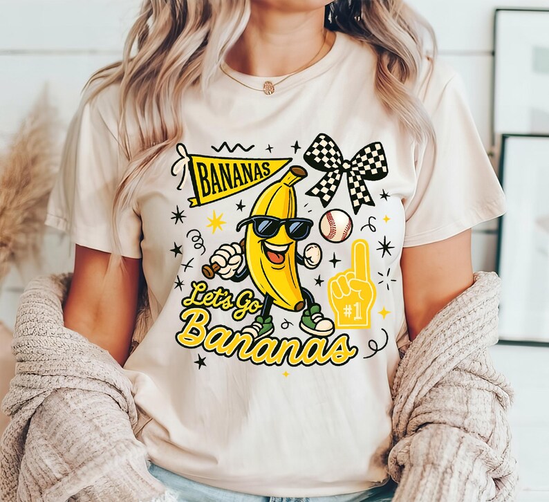 May include: Cream-colored t-shirt featuring a cartoon banana wearing sunglasses, holding a baseball bat, and wearing sneakers. The shirt has the text "Bananas" and "Let's Go Bananas" in yellow and black, along with baseball-themed graphics.
