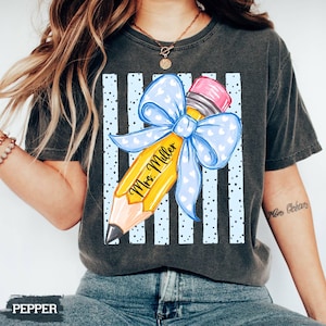 May include: A charcoal gray t-shirt featuring a graphic of a yellow pencil with a pink eraser and a large light blue bow with white hearts. The pencil is printed with the text Mrs. Miller, set against a background of vertical light blue stripes.