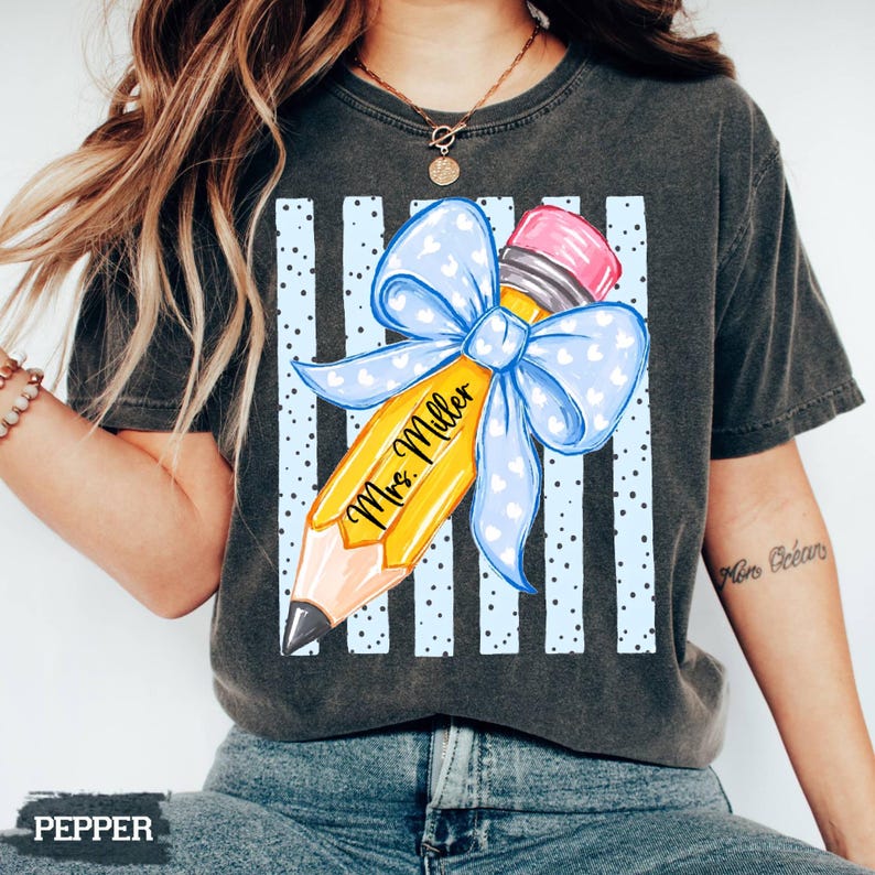 May include: A charcoal gray t-shirt featuring a graphic of a yellow pencil with a pink eraser and a large light blue bow with white hearts. The pencil is printed with the text Mrs. Miller, set against a background of vertical light blue stripes.