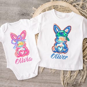 May include: Two white t-shirts with colorful bunny illustrations. The bunny on the left has a pink bow and the bunny on the right has a green baseball cap. The shirts are personalized with the names "Olivia" and "Oliver".
