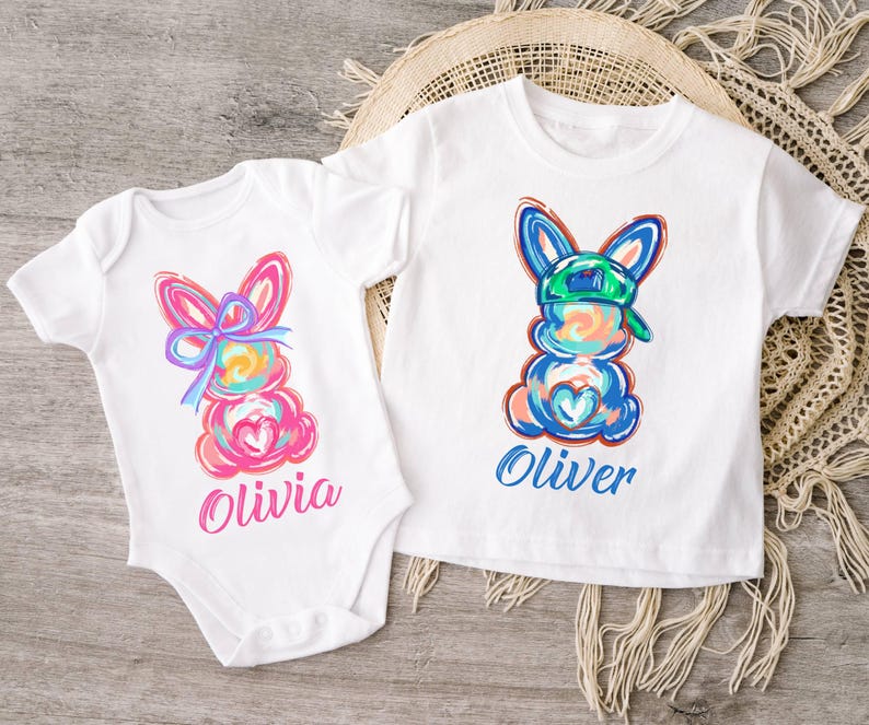 May include: Two white t-shirts with colorful bunny illustrations. The bunny on the left has a pink bow and the bunny on the right has a green baseball cap. The shirts are personalized with the names "Olivia" and "Oliver".