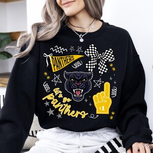 May include: Black sweatshirt featuring a panther graphic with the text "Panthers" on a pennant and "Let's Go Panthers". The design includes a checkered bow, a foam finger, and star accents. The sweatshirt is a crew neck.