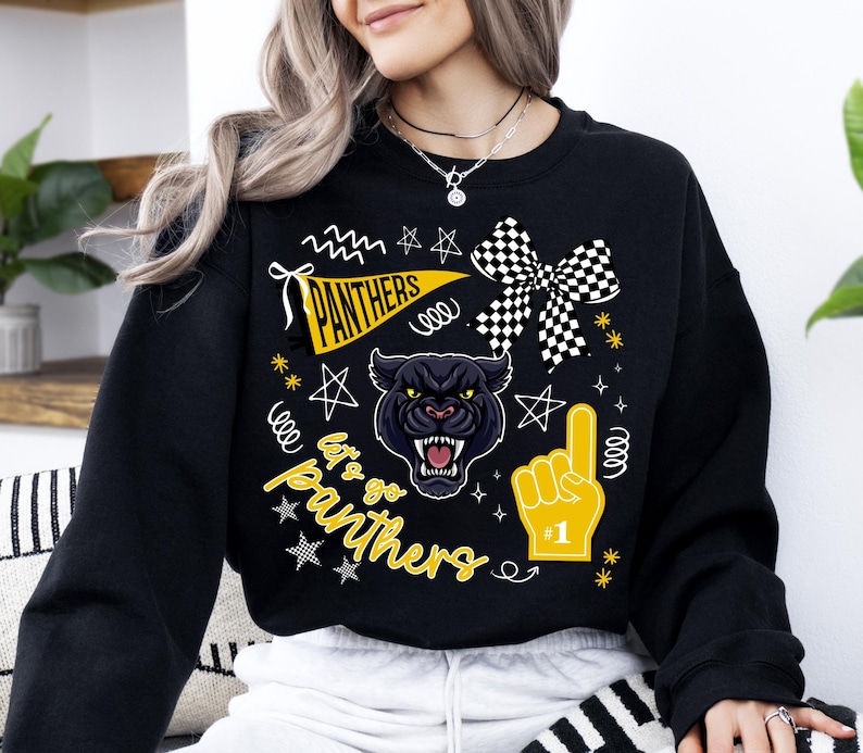 May include: Black sweatshirt featuring a panther graphic with the text "Panthers" on a pennant and "Let's Go Panthers". The design includes a checkered bow, a foam finger, and star accents. The sweatshirt is a crew neck.